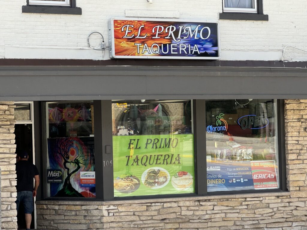 Storefront of "El Primo Taqueria" with a brightly colored sign, menu display in the window, and a man entering through a door on the left.