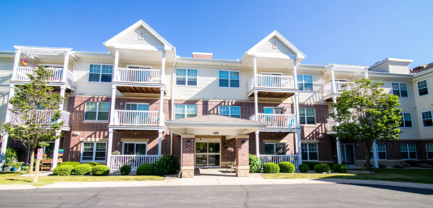 Three-story apartment building with brick and siding exterior, multiple balconies, and a covered entrance, surrounded by small trees and bushes on a sunny day.