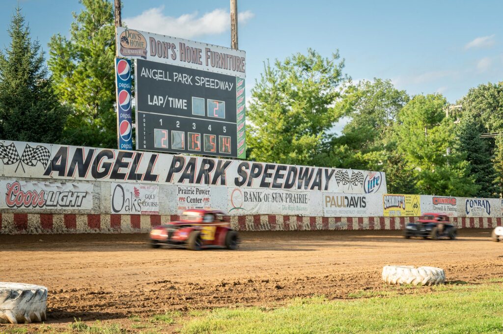 Two race cars speed along a dirt track at Angell Park Speedway, with a scoreboard and sponsor advertisements visible in the background.