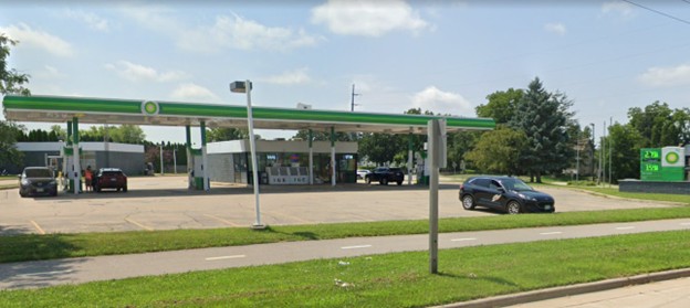A BP gas station with two cars parked by the pumps and another car driving on a nearby road, under a partly cloudy sky.