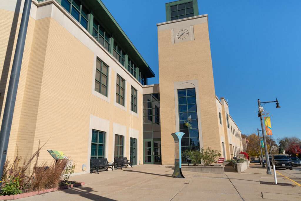 A modern beige brick building with a clock tower, multiple windows, and benches by the entrance on a clear, sunny day.