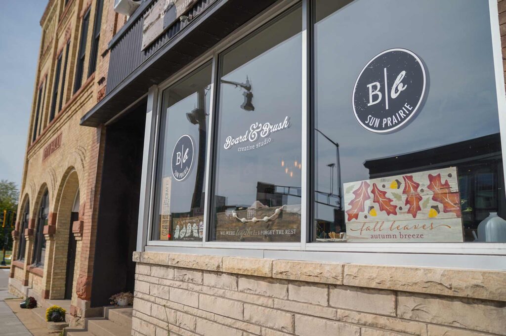 Street-level view of a brick building with large windows displaying “B&Co Sun Prairie” branding and autumn-themed decorations.