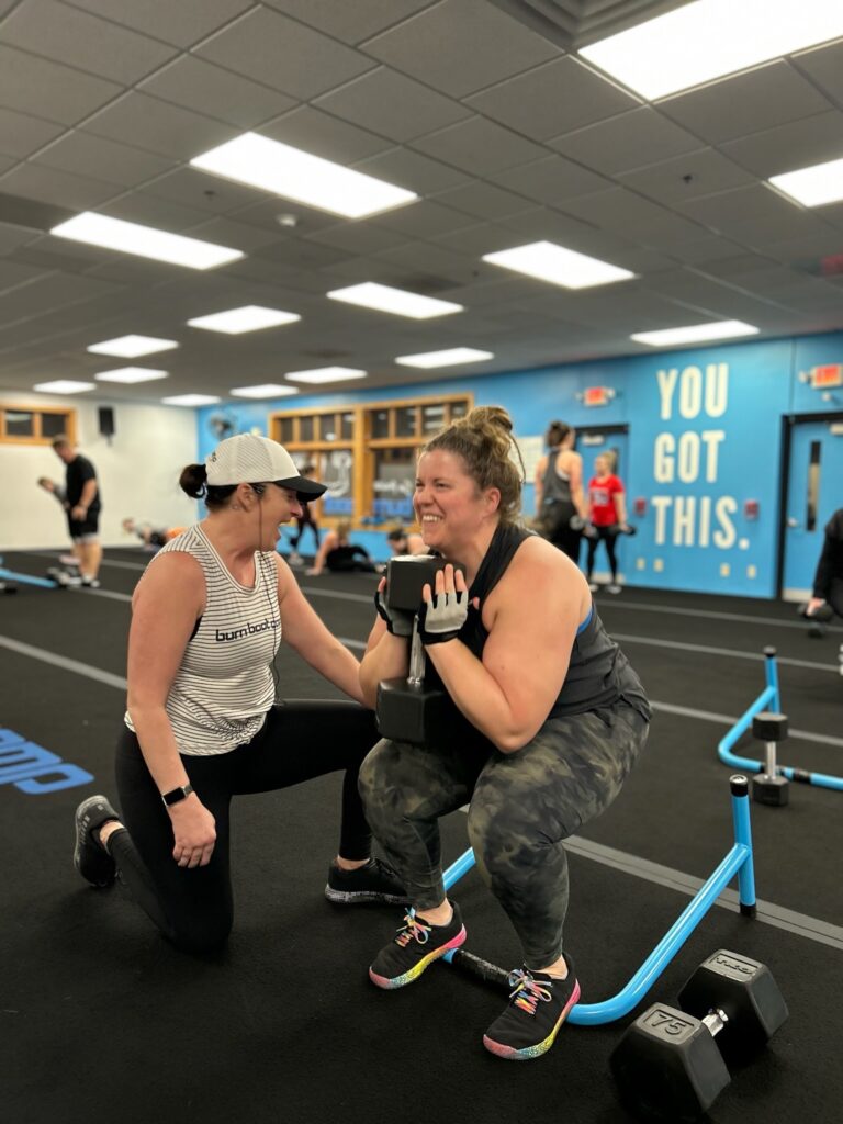 A woman performs a squat with dumbbells while another woman coaches her in a gym. Others exercise in the background; a wall reads, "YOU GOT THIS.