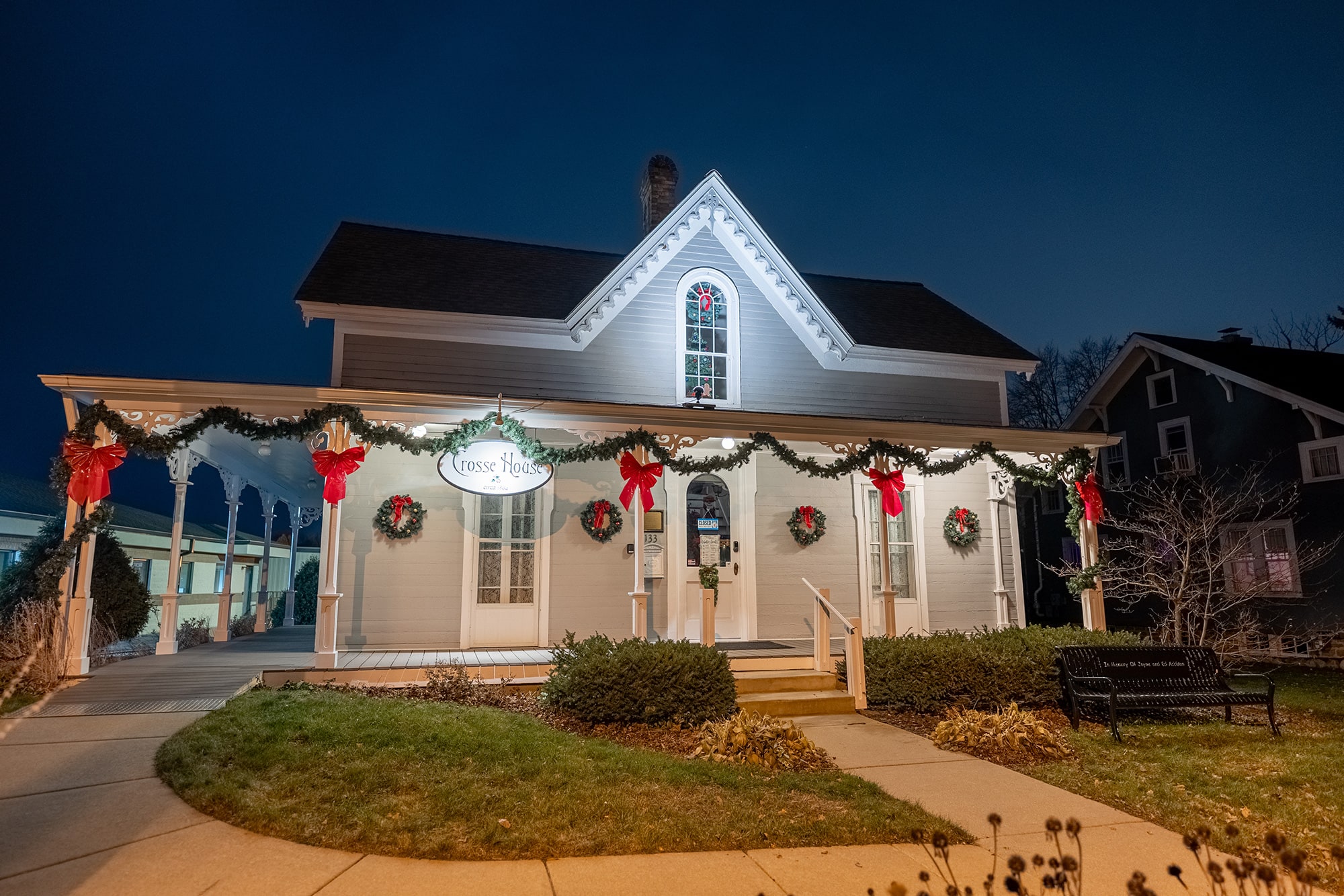 A white, two-story house decorated with wreaths, garlands, and red bows for the holidays is lit up at night. A sign above the door reads "Cross House.