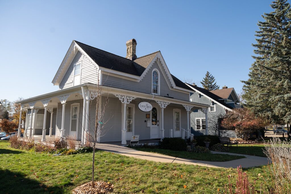 A white Victorian-style house with decorative trim, a wraparound porch, and a sign reading "Open House" on the front, surrounded by grass and trees on a clear day.
