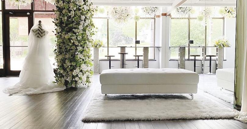 A bride in a wedding gown stands by a large floral arrangement near tall windows in a bright, modern lounge with white furniture and dark wood floors.