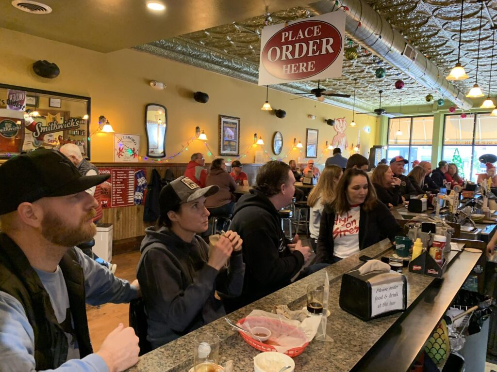 People sit at a crowded bar, eating and drinking. A "Place Order Here" sign hangs above the counter. The atmosphere appears casual and lively.