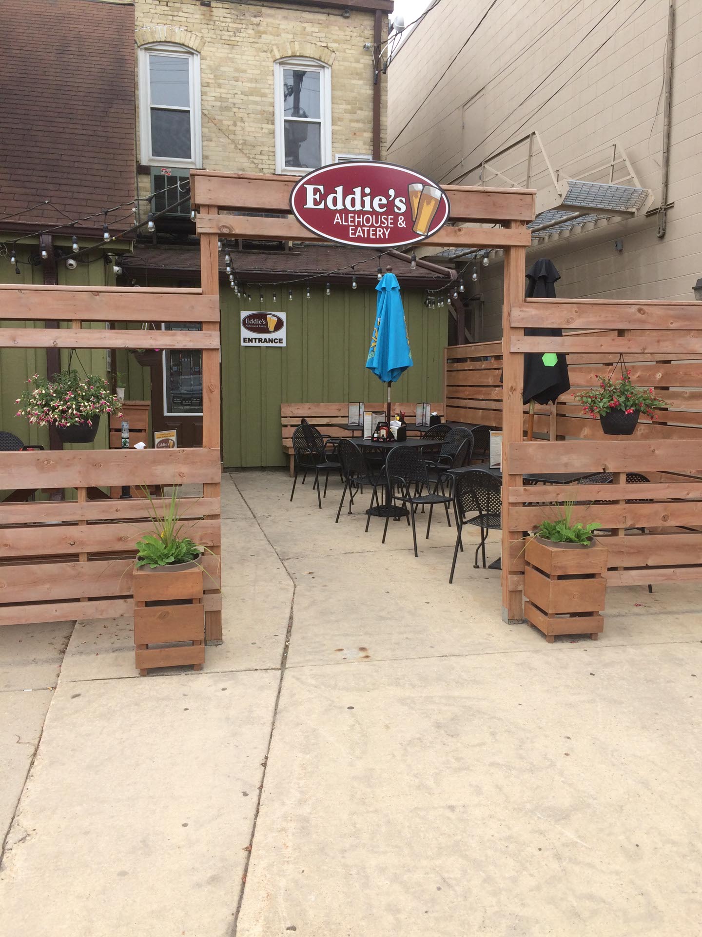 Outdoor patio entrance to Eddie's Alehouse & Eatery with wooden fencing, tables, chairs, and potted plants visible.