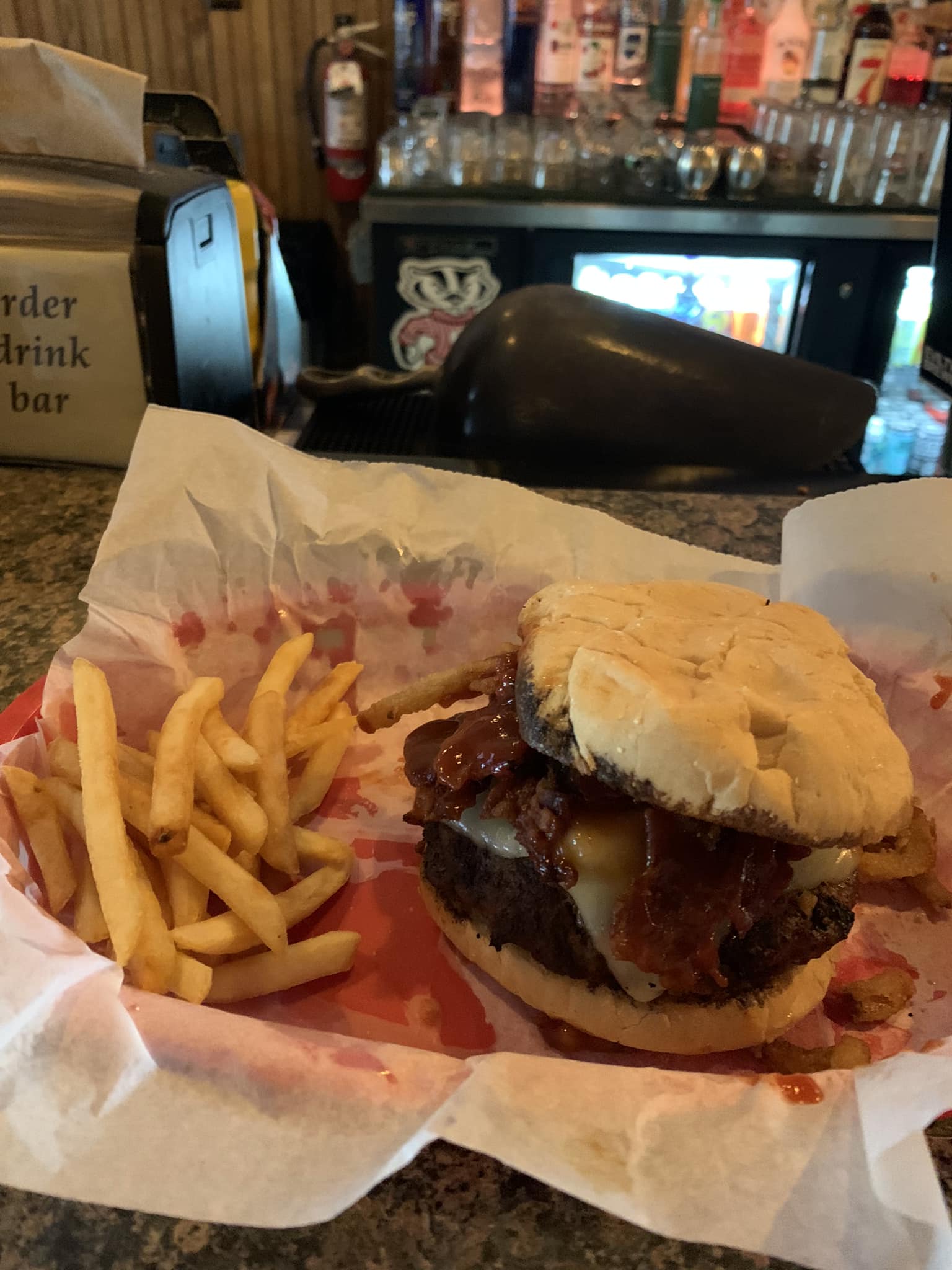 A cheeseburger with bacon, onion rings, and barbecue sauce is served with a side of French fries on a tray lined with wax paper at a bar counter.
