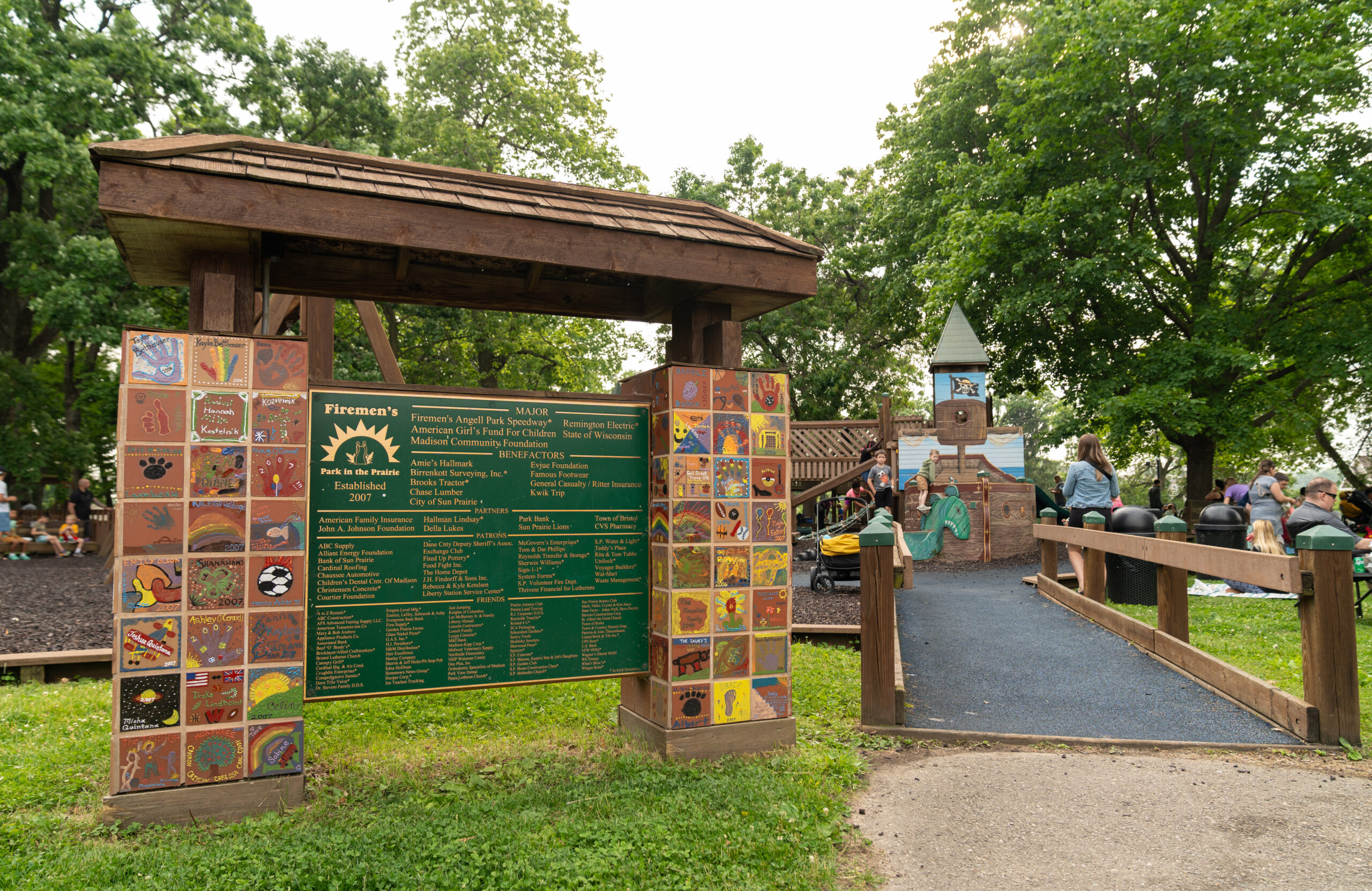 A large wooden sign with names and colorful tiles stands at the entrance to a playground, with trees, children, and adults visible in the background.