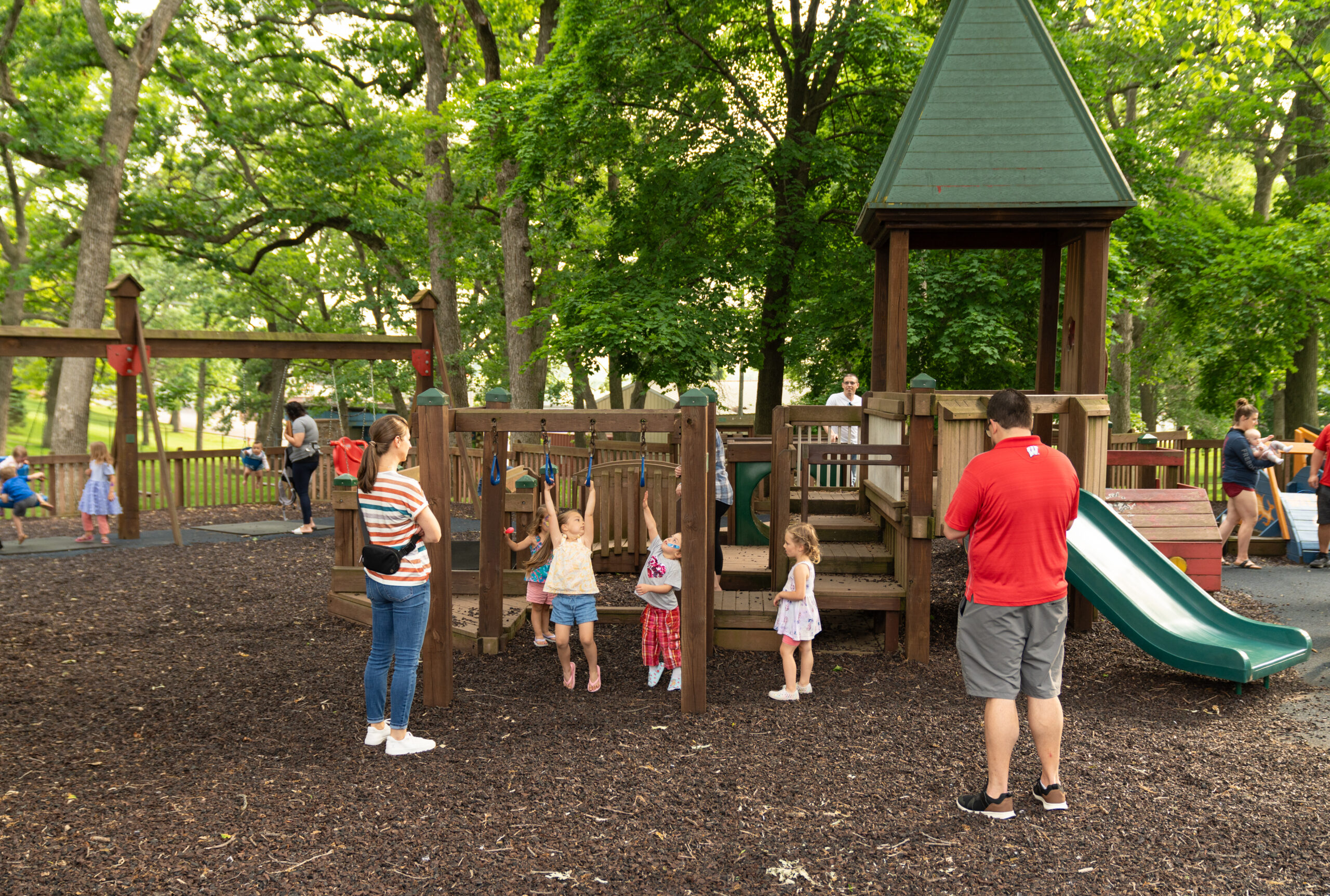 Children play on playground equipment while adults stand nearby in a park surrounded by trees. Some kids use a slide, others play beneath a structure.