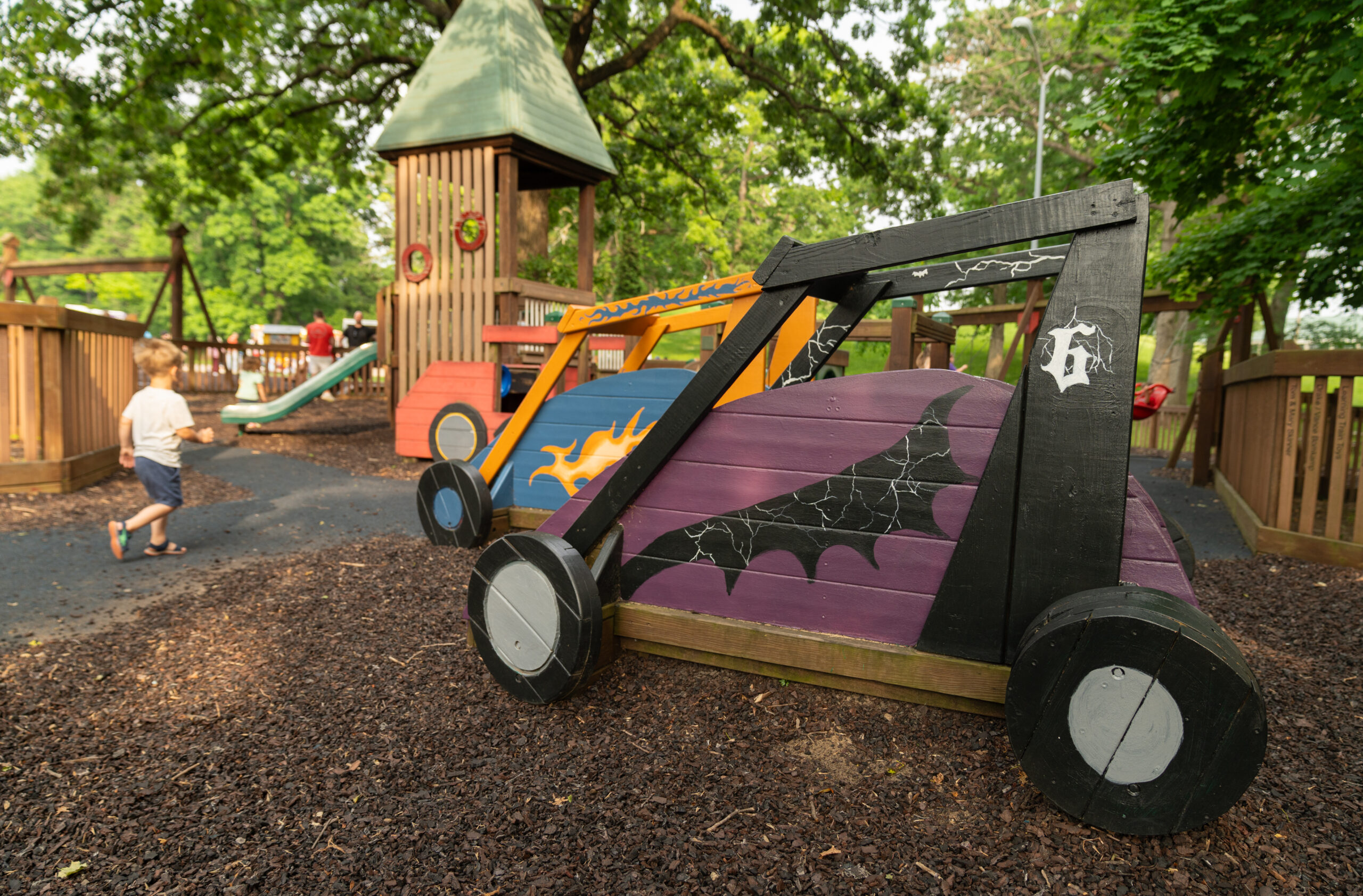 A young child walks in a playground with wooden play structures, including colorful car-shaped installations, surrounded by trees.
