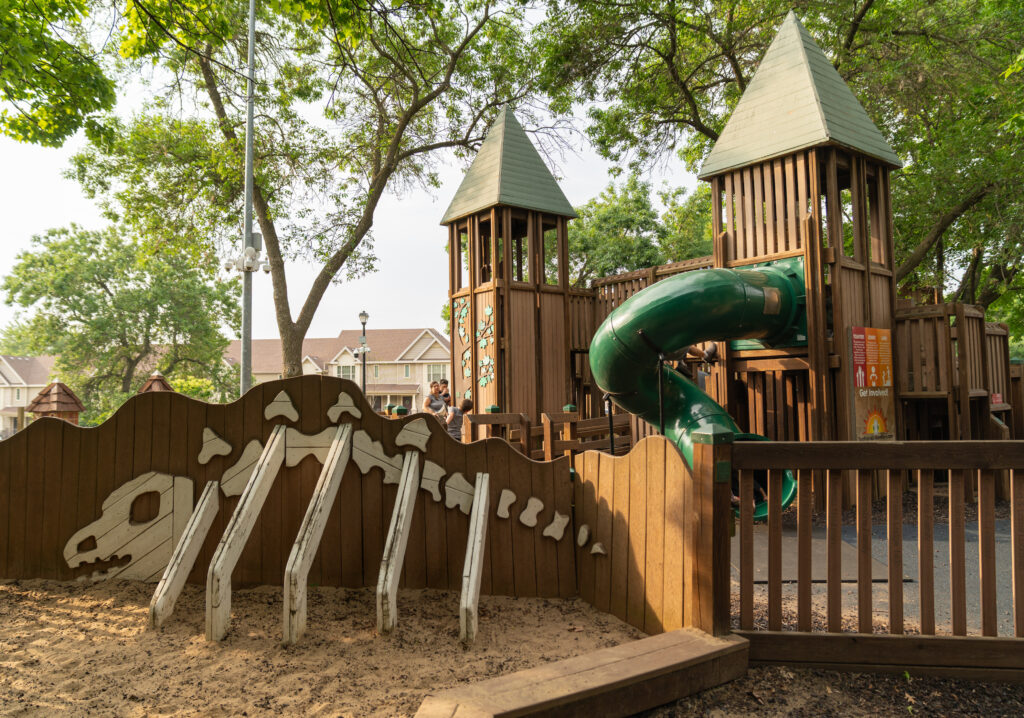 Wooden playground with towers, a green spiral slide, and a dinosaur skeleton model partially buried in a sand area beneath trees.