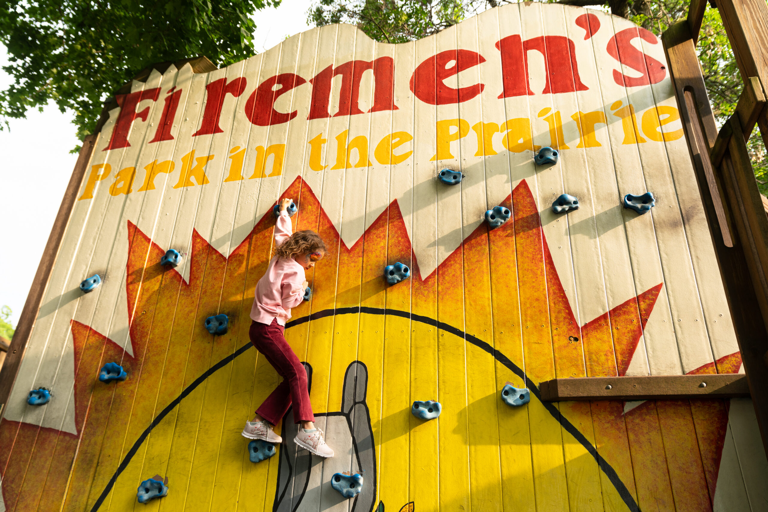 A child climbs a rock wall at Firemen's Park in the Prairie, with colorful text and sunburst graphics painted on the wooden structure.