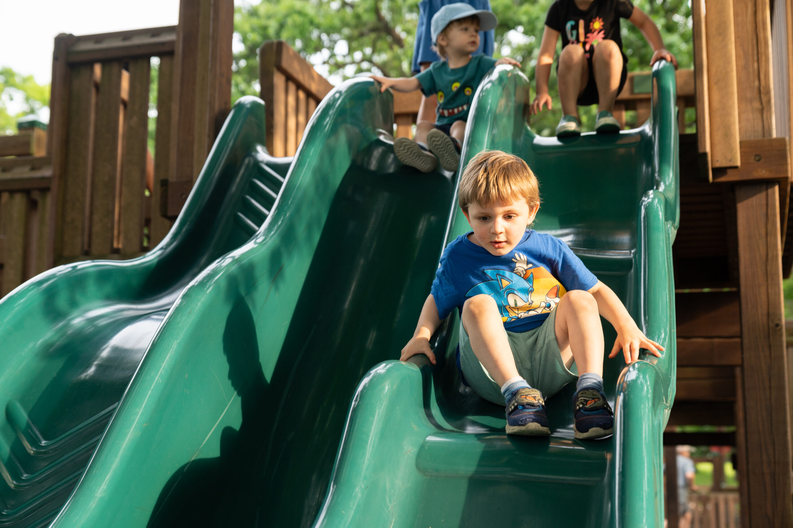 A young boy in a blue shirt slides down a green playground slide, with two other children waiting at the top in the background.