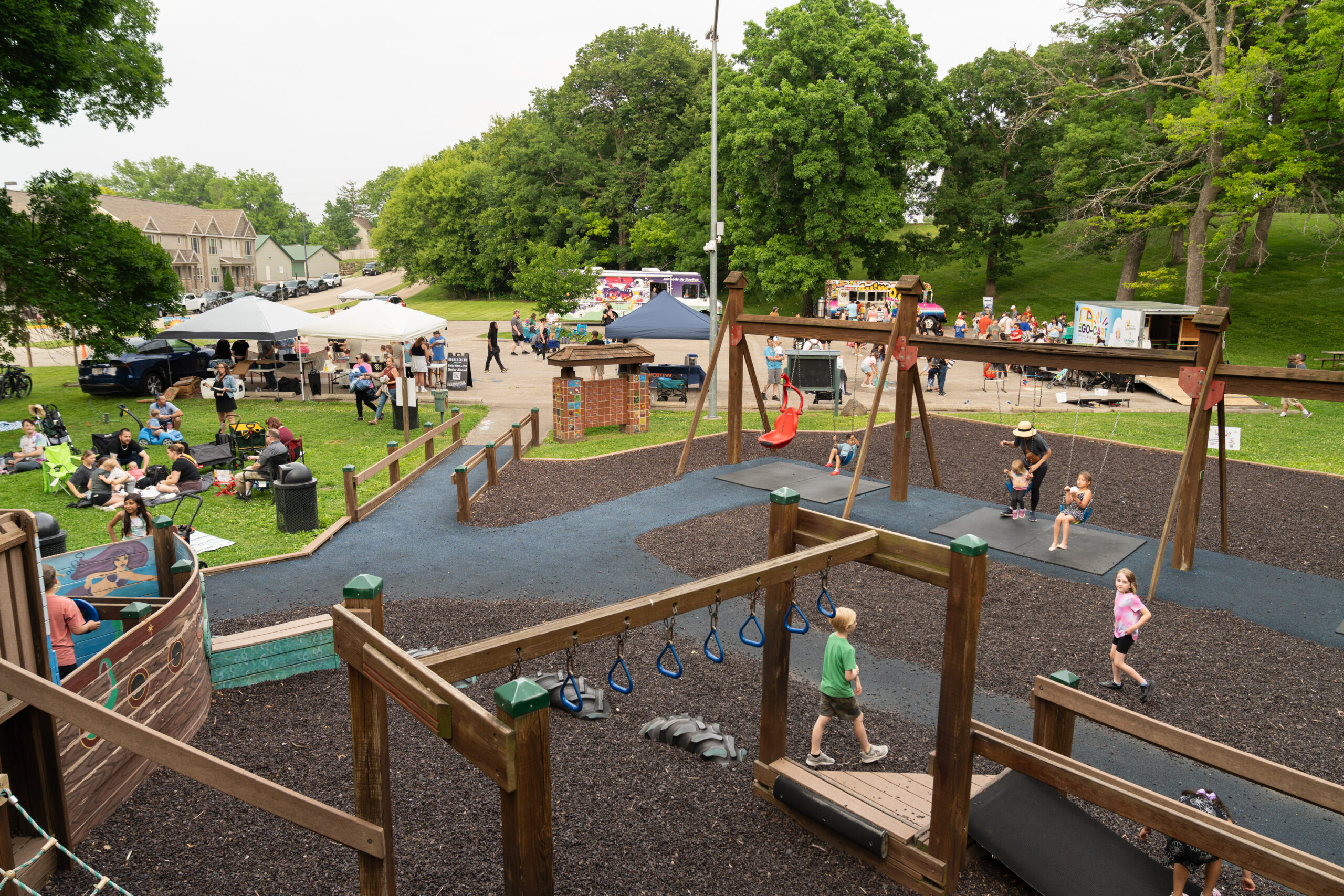 Children play on a playground while adults gather nearby at an outdoor event with food trucks and tents in a park setting.