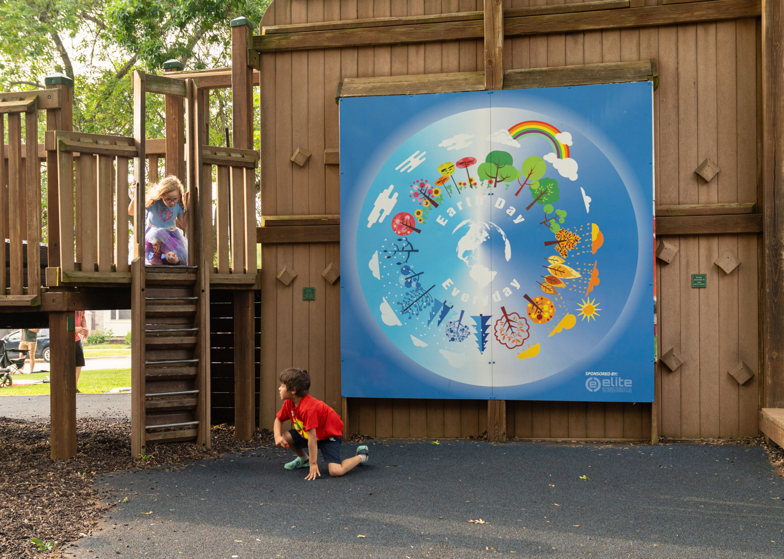 Two children play near a large, colorful mural on a wooden playground structure. The mural features trees, clouds, and weather symbols in a circular design.