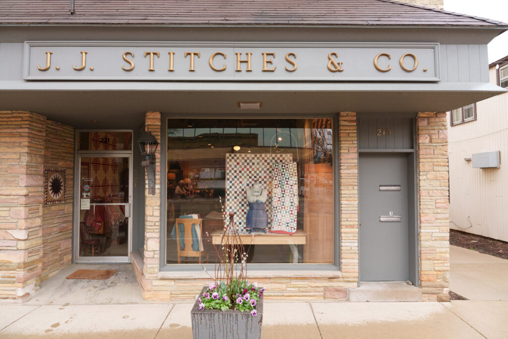 The storefront of J.J. Stitches & Co. features a large display window showing a quilt and a mannequin, with the shop’s name in bold letters above the entrance.