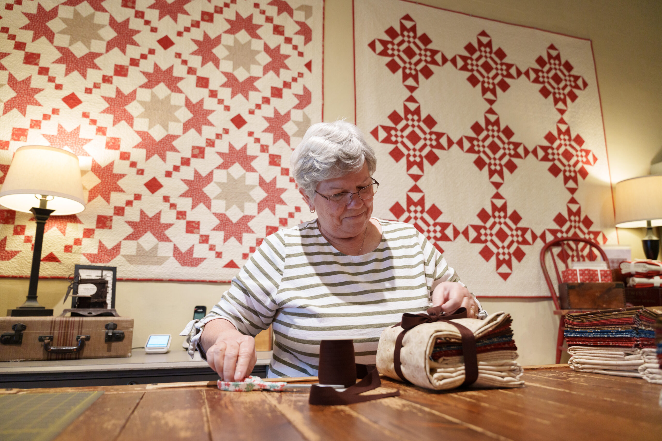 An older woman in a striped shirt folds and ties a stack of fabric at a wooden table, with red and white patchwork quilts hanging on the wall behind her.