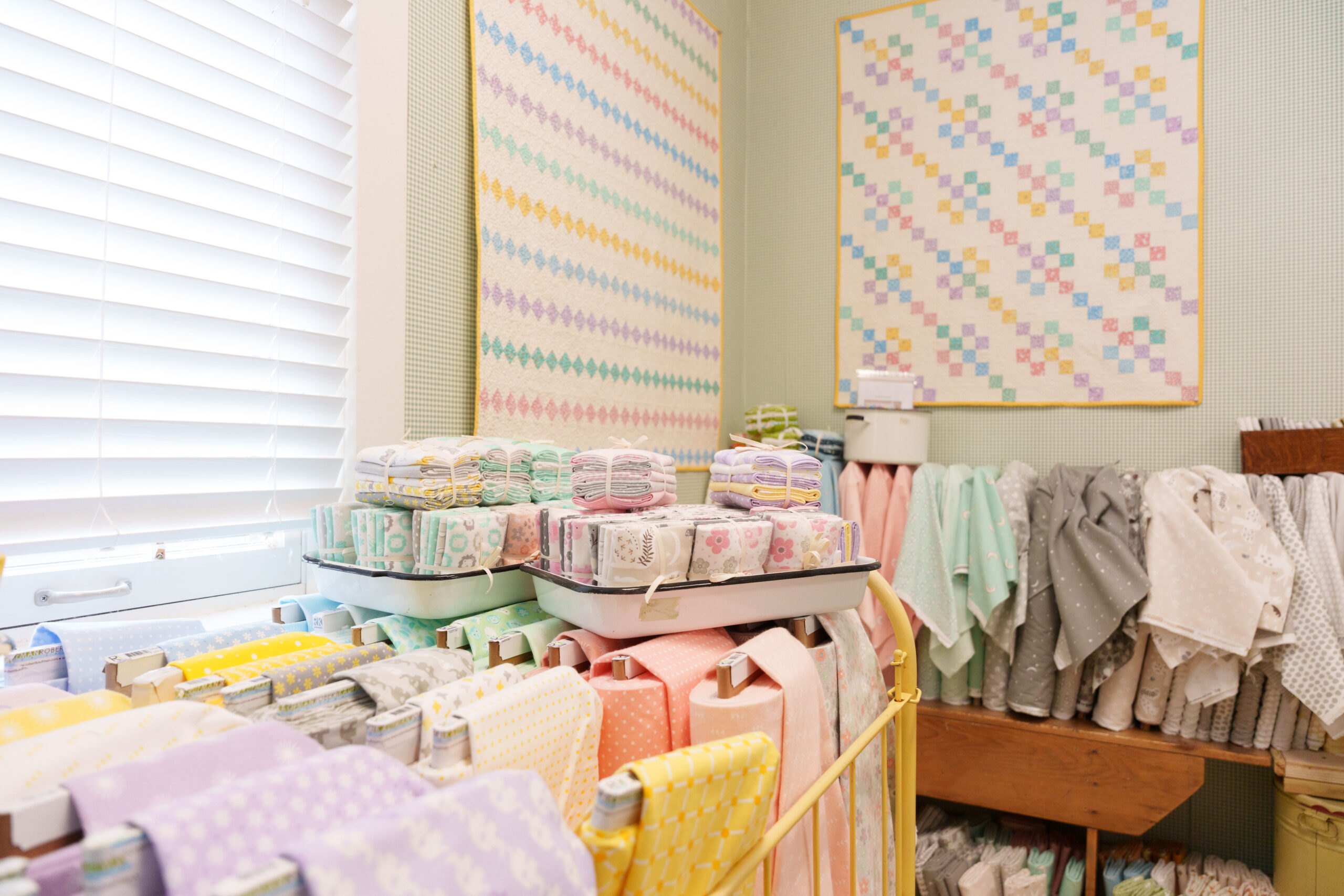 Shelves and racks display neatly folded and rolled pastel-colored fabrics in a well-lit room, with patterned quilts hanging on the walls.