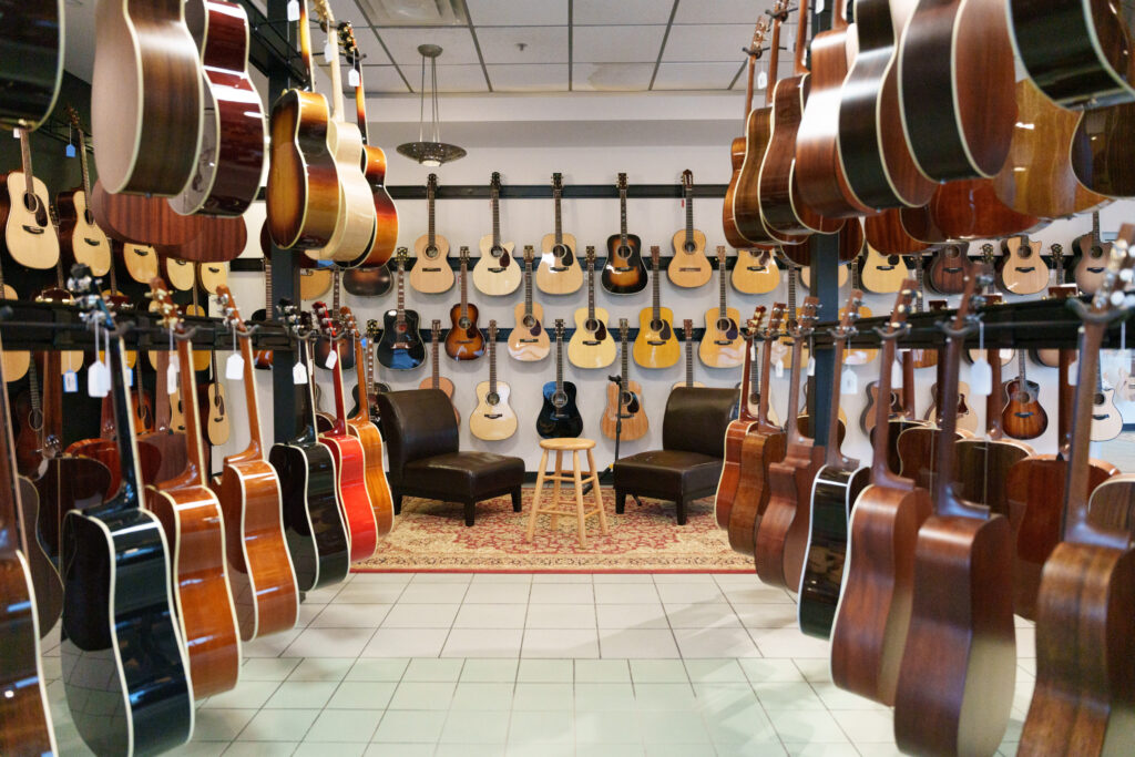 Rows of acoustic guitars hang on the walls and racks in a music store, with two chairs and a small table placed on a rug in the center of the room.