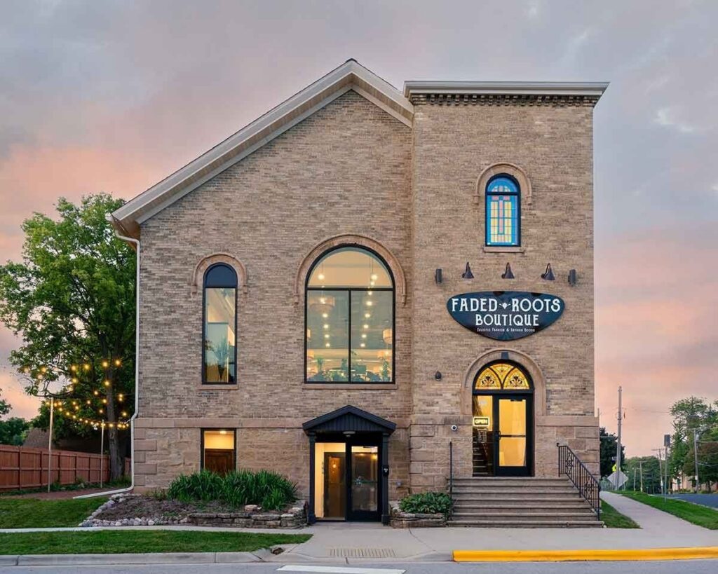 A two-story brick building with large arched windows and a sign reading "Faded Roots Boutique" above the entrance; exterior lights and greenery are visible.