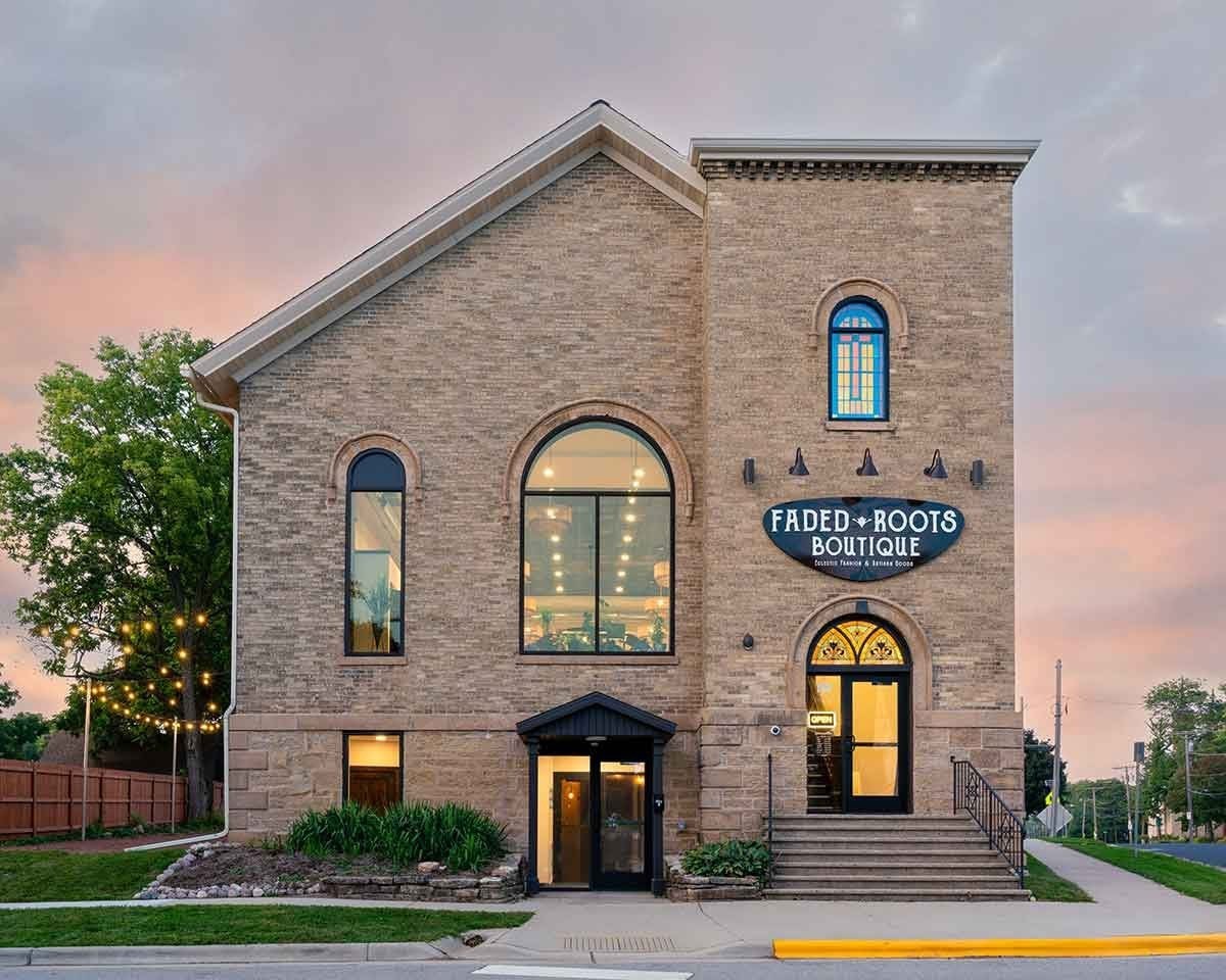 A two-story brick building with large arched windows and a sign reading "Faded Roots Boutique" above the entrance; exterior lights and greenery are visible.
