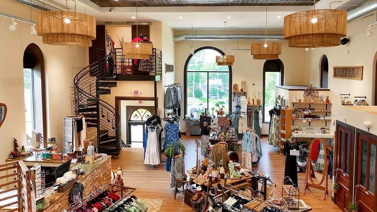 A boutique store interior with clothing racks, shelves of accessories, mannequins, and large arched windows, featuring wooden floors and spiral staircase.