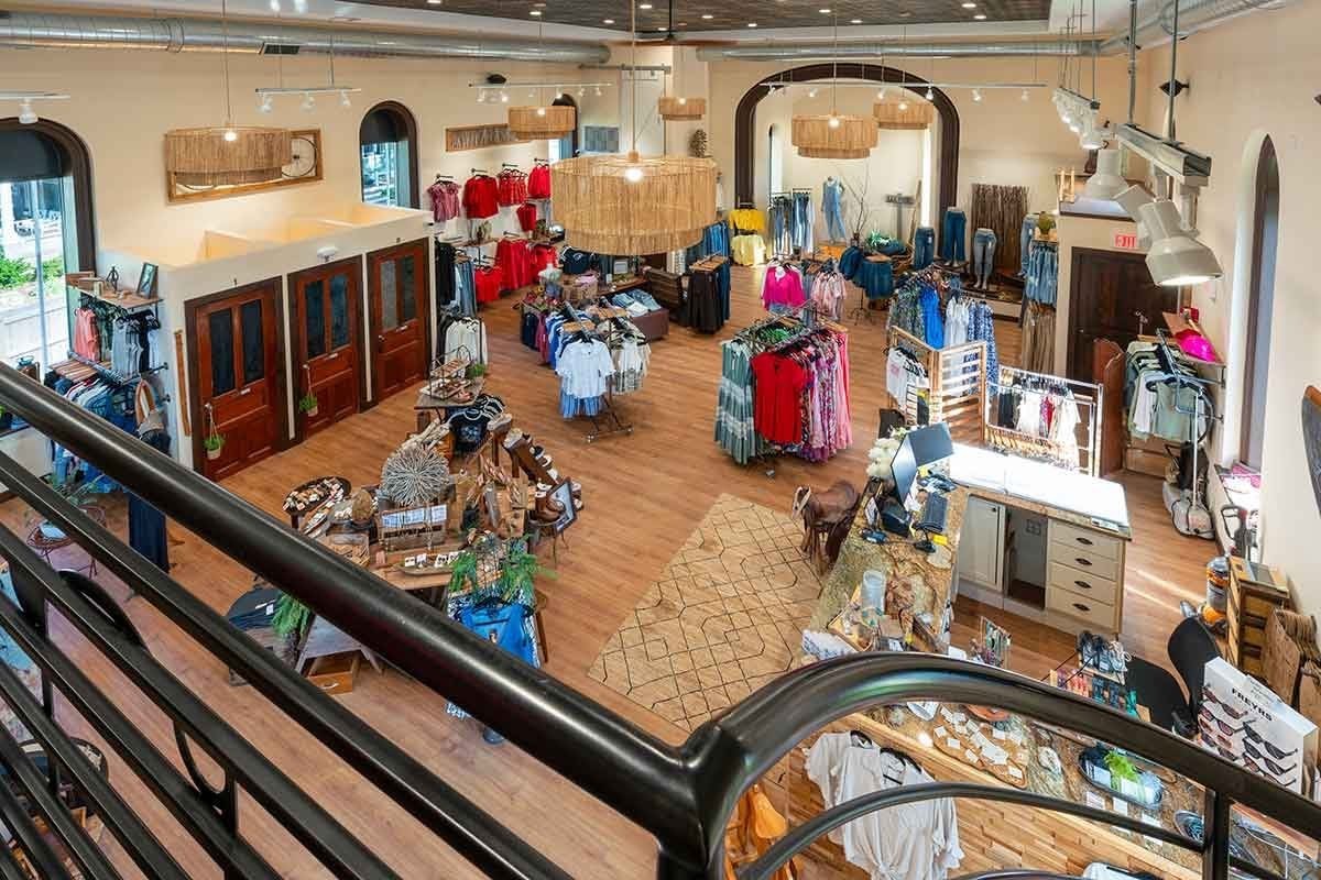 A clothing and gift boutique with wooden floors, racks of apparel, tables of accessories, and natural light from arched windows, viewed from an upper-level railing.