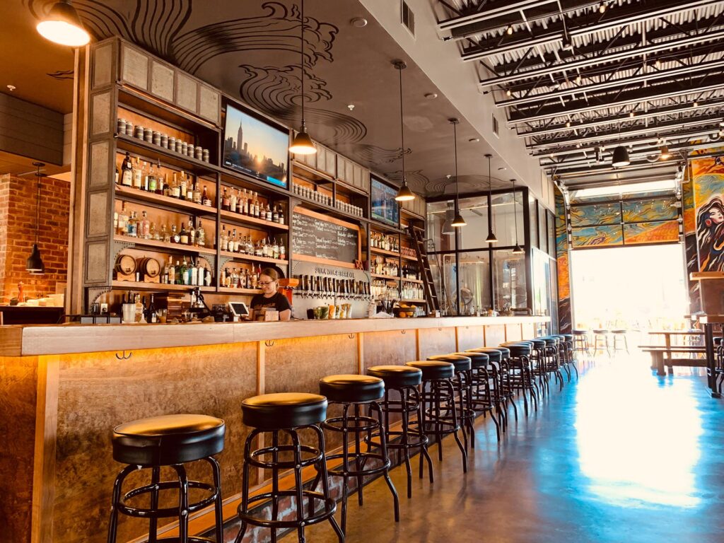 A modern bar with a long counter, high stools, shelves stocked with bottles, and a bartender standing behind the bar in a brightly lit, stylish interior.