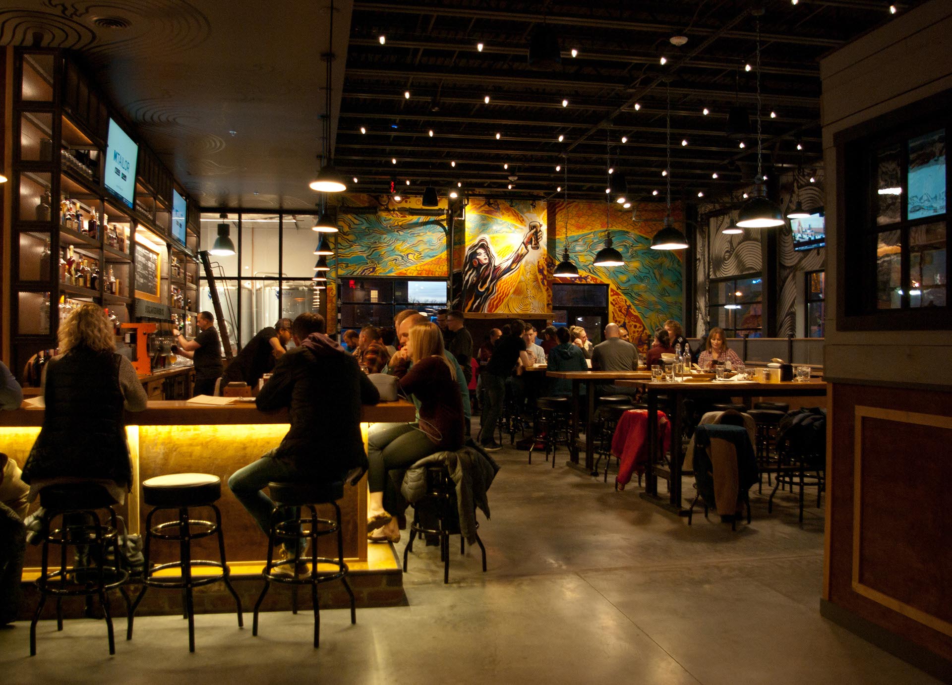 A modern bar with people sitting at the counter and tables, under string lights, with a colorful mural on the back wall.
