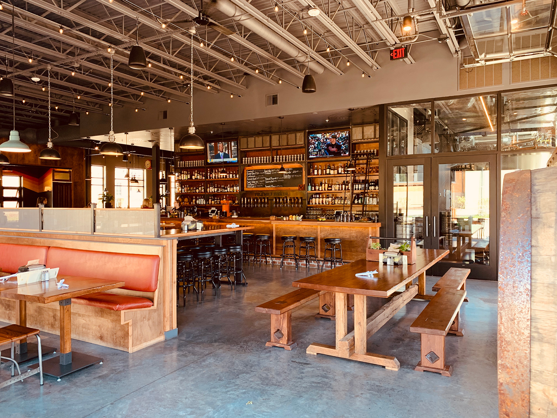 Spacious restaurant interior with wooden tables, red booth seating, bar area, and shelves stocked with bottles; TVs mounted above the bar. Natural light enters through large windows.