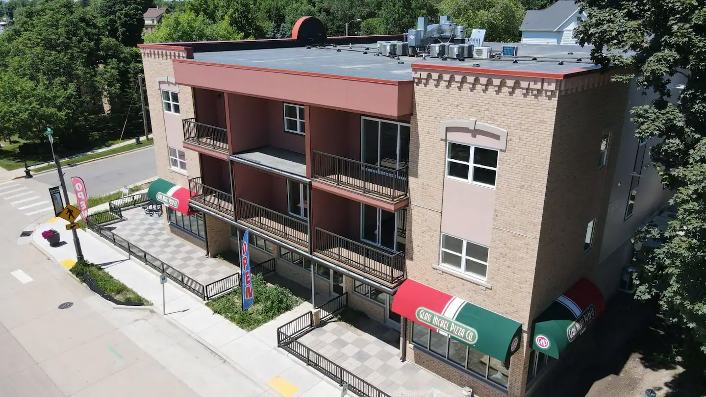 A three-story brick building with multiple balconies, red and green awnings, and a pizza restaurant on the ground floor, located on a tree-lined street corner.
