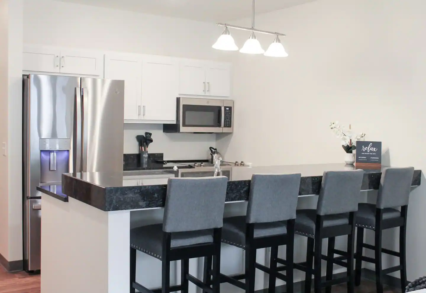 Modern kitchen with stainless steel appliances, white cabinets, black countertops, and four gray upholstered barstools at a breakfast bar. A small plant sits on the counter.