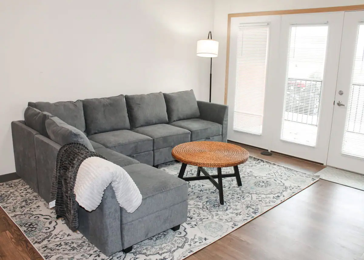 A modern living room with a gray sectional sofa, a round wicker coffee table, a floor lamp, and large windows with blinds letting in natural light.