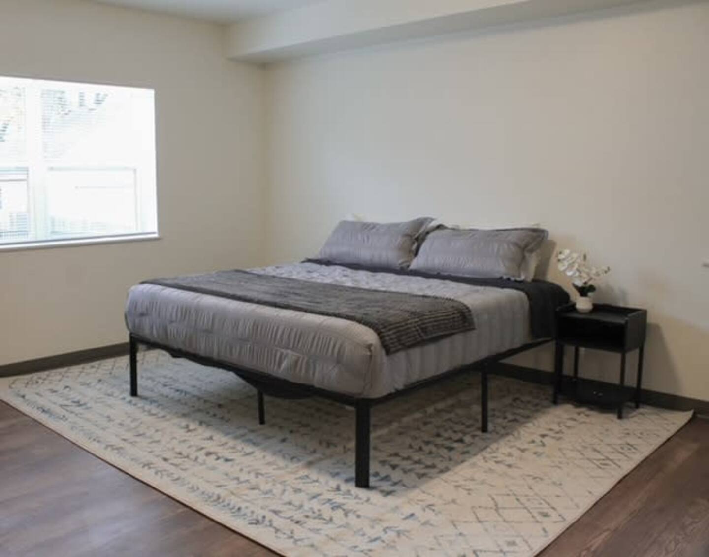 A neatly made bed with gray bedding sits on a patterned rug in a minimally furnished bedroom with wood floors, a window, and a black nightstand holding a small decorative item.