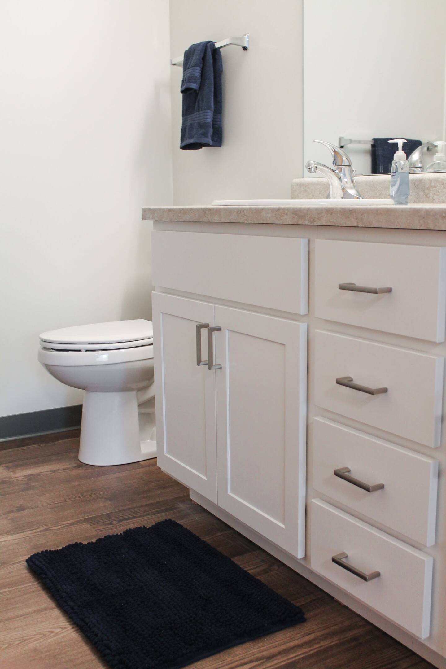 A modern bathroom with white cabinets, a countertop sink, a toilet, a dark blue towel on a rack, and a matching dark blue floor mat on wooden flooring.
