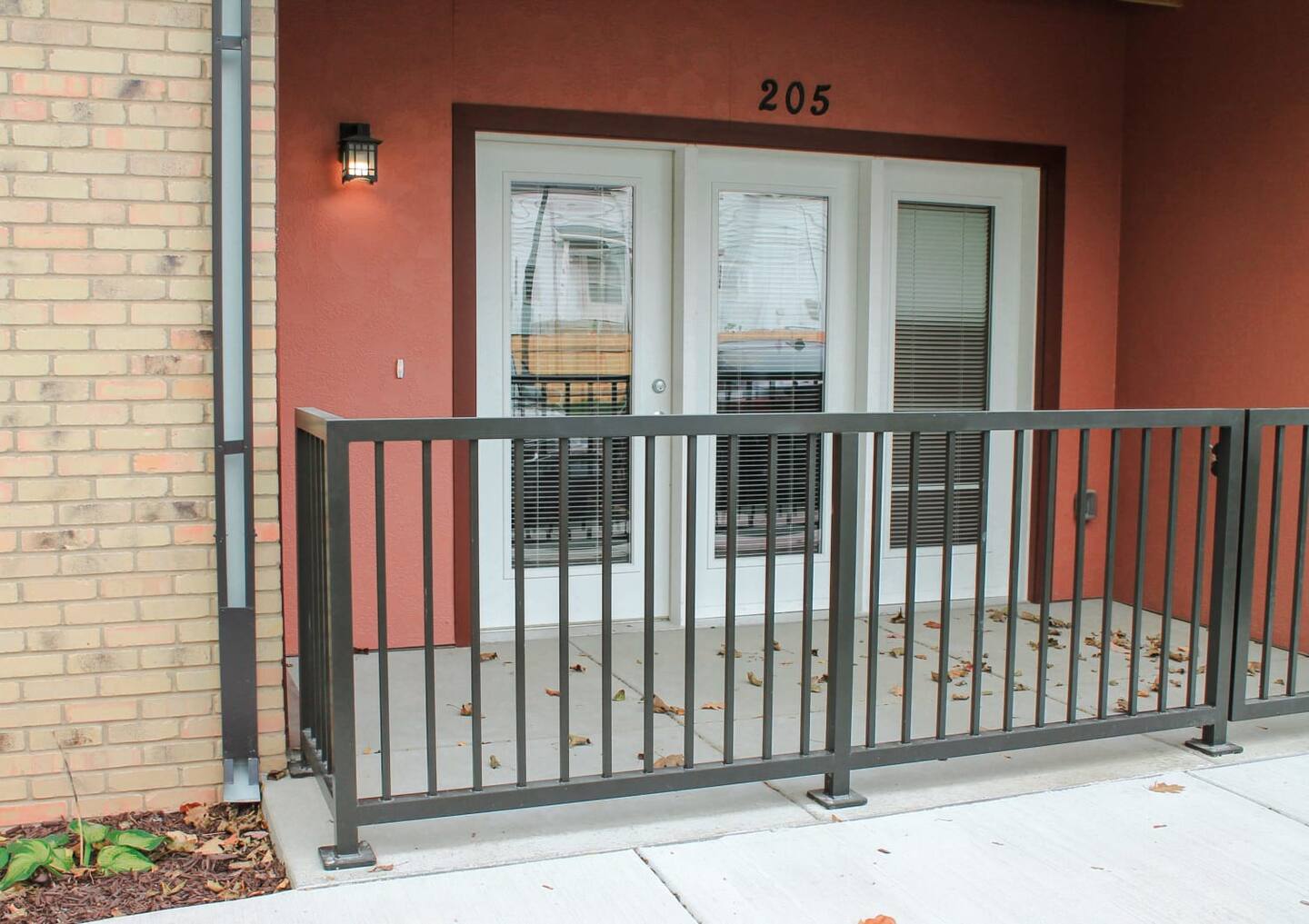 A patio with a black metal railing, light brown brick wall on the left, two white double doors with blinds, and the number 205 above the doors. Some leaves are scattered on the ground.