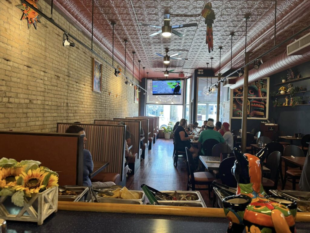 A restaurant interior with brick walls, booth and table seating, a TV on the back wall, and decorative items including a ceramic rooster and sunflowers in the foreground.