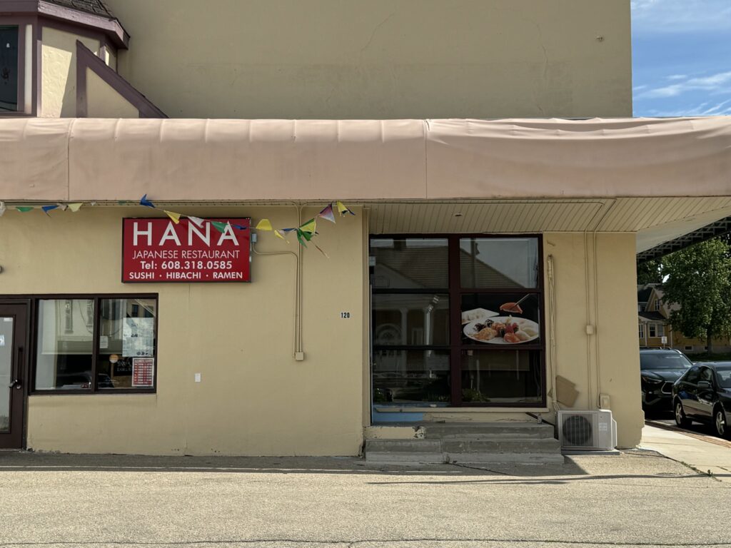 Front view of Hana Japanese Restaurant with a red sign, tan exterior, a food poster by the entrance, and colorful flags hanging above.