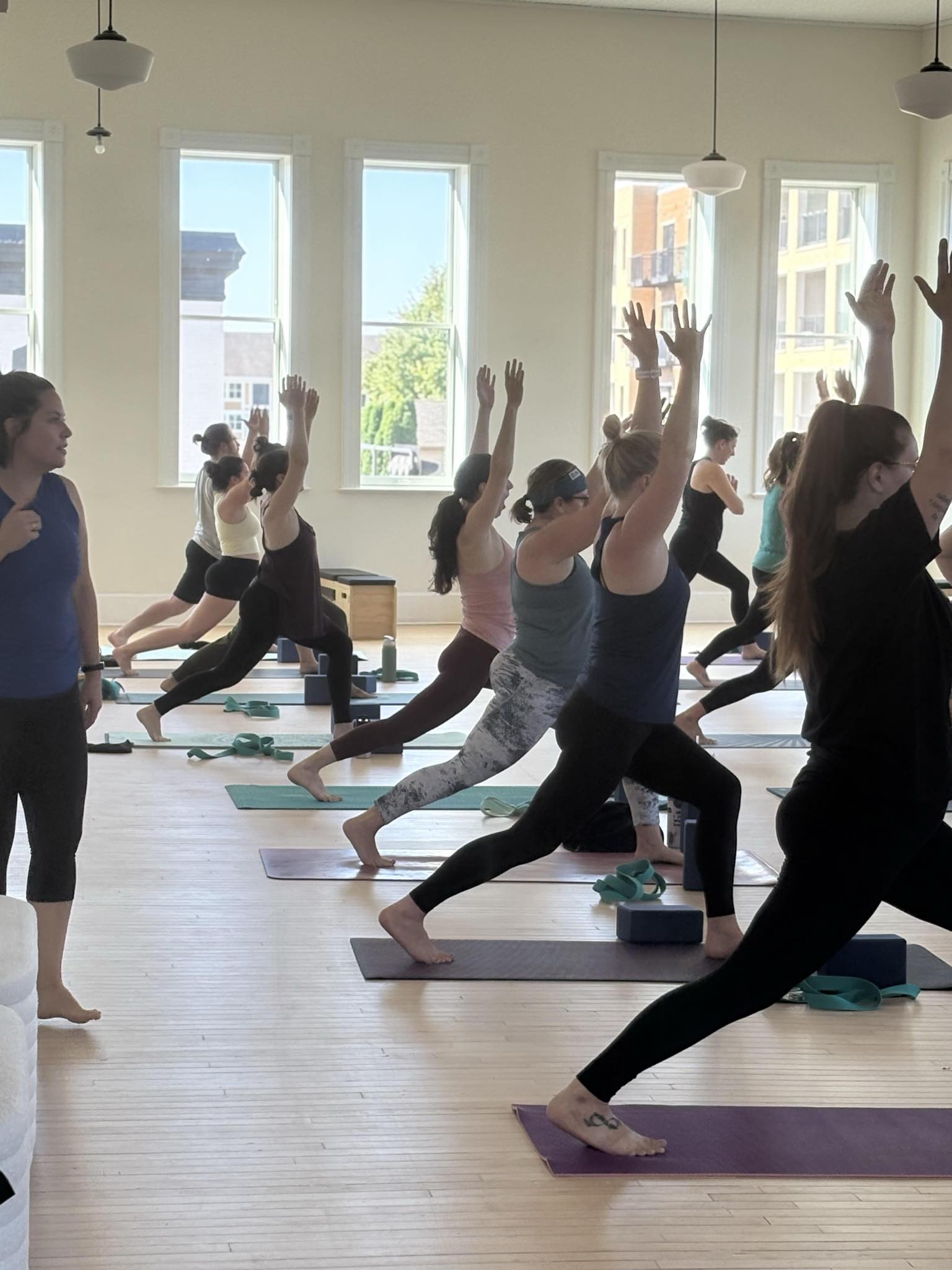 A group of people practice yoga in a bright studio, performing a standing pose with arms raised, while an instructor observes.