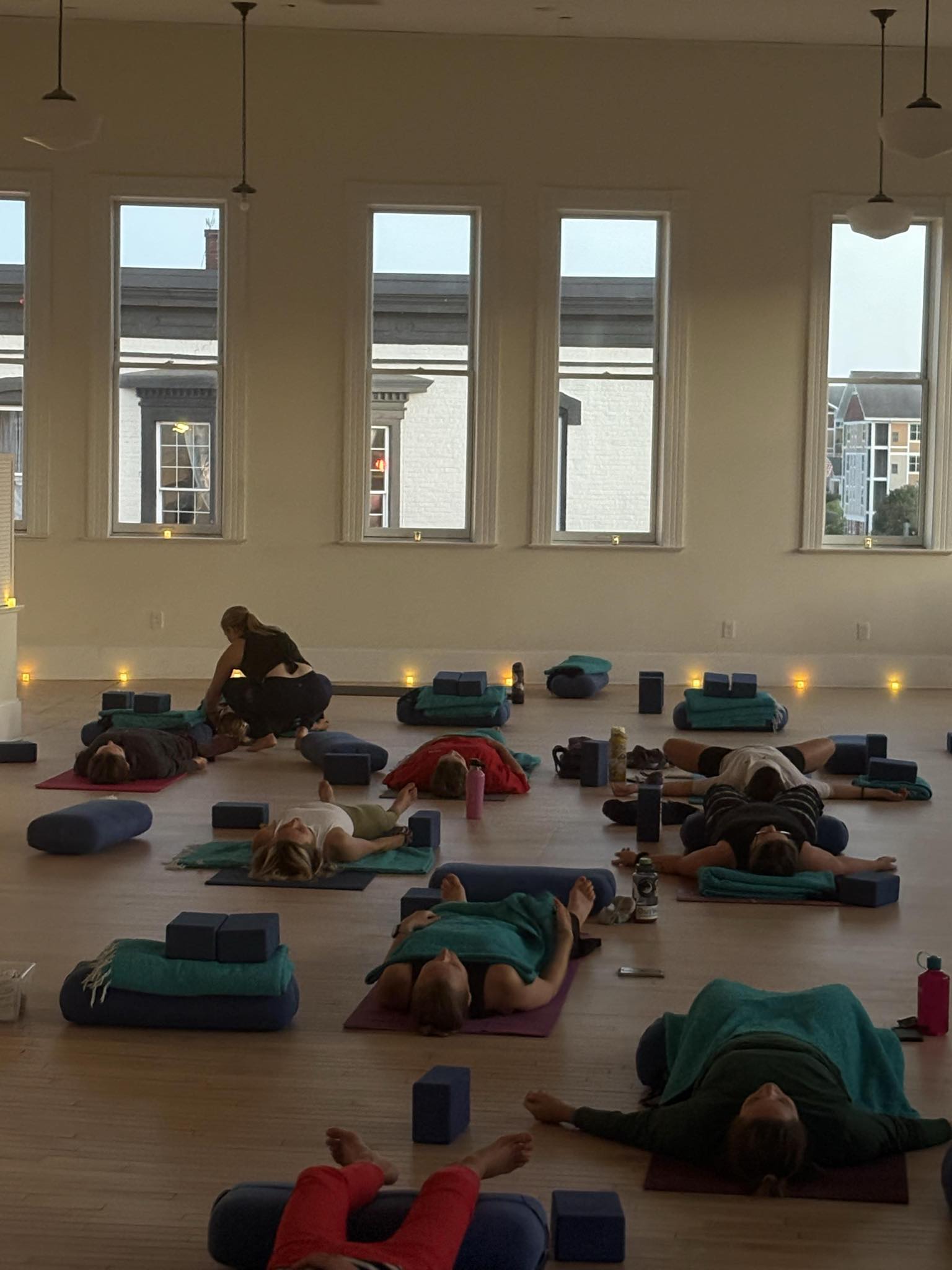 A group of people lying on yoga mats in a studio, with props such as blocks and bolsters, while an instructor assists one participant.