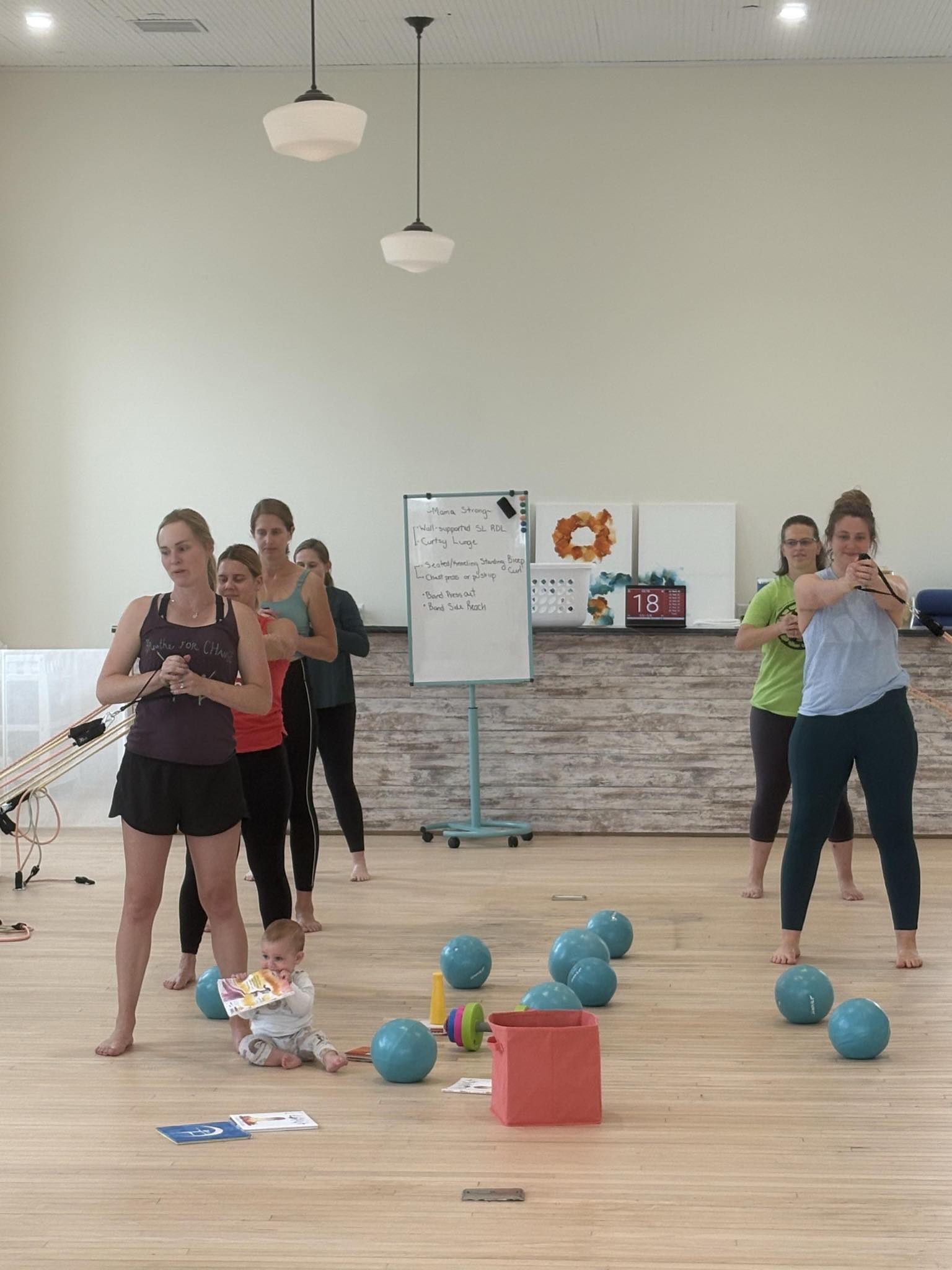 A group of women participate in a fitness class in a studio. A baby sits on the floor with toys and books among exercise balls and props. A whiteboard is visible in the background.