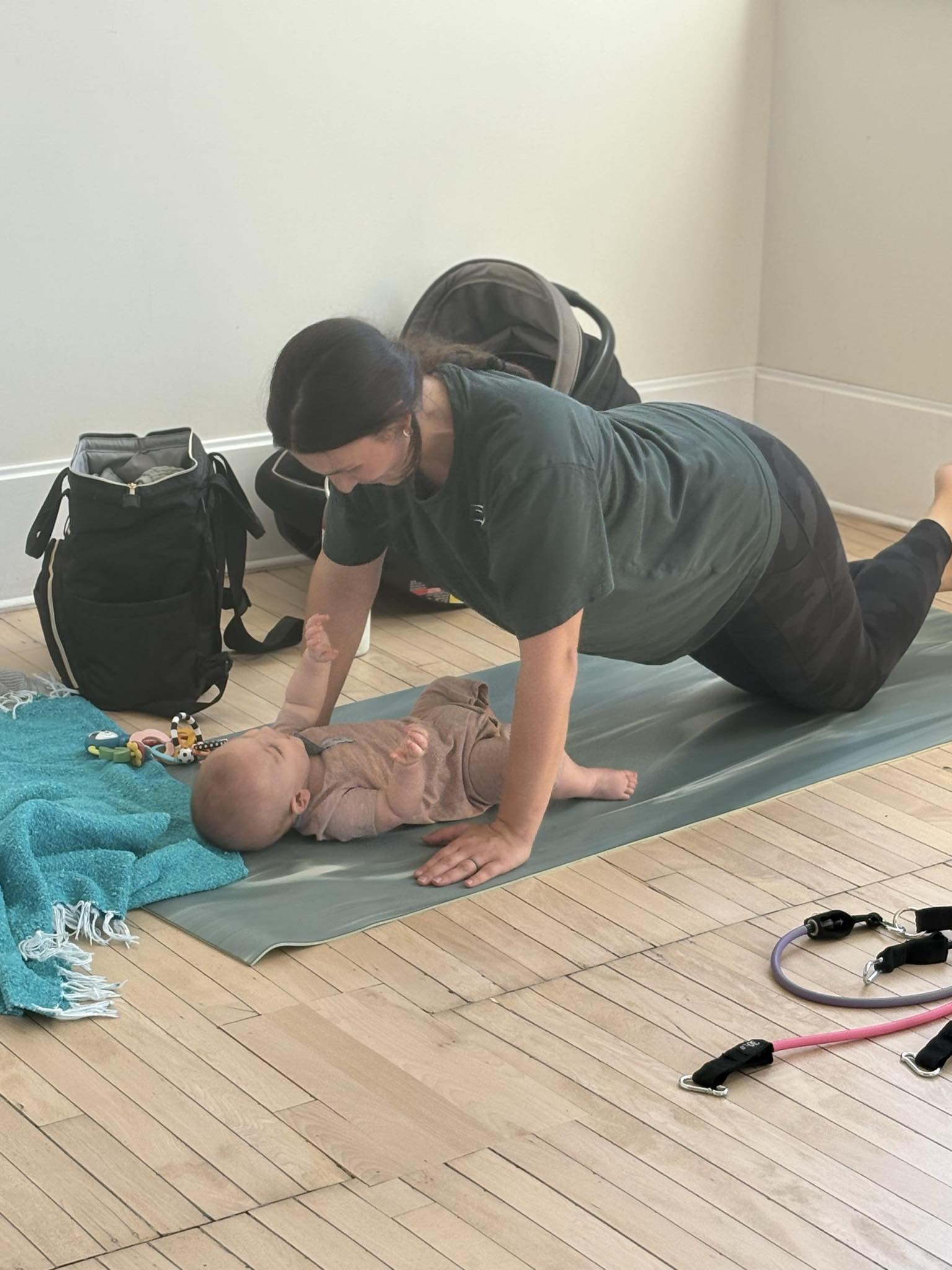 A woman does a modified push-up on a yoga mat while facing a baby lying in front of her. Bags, a blue blanket, and exercise bands are nearby on a wooden floor.