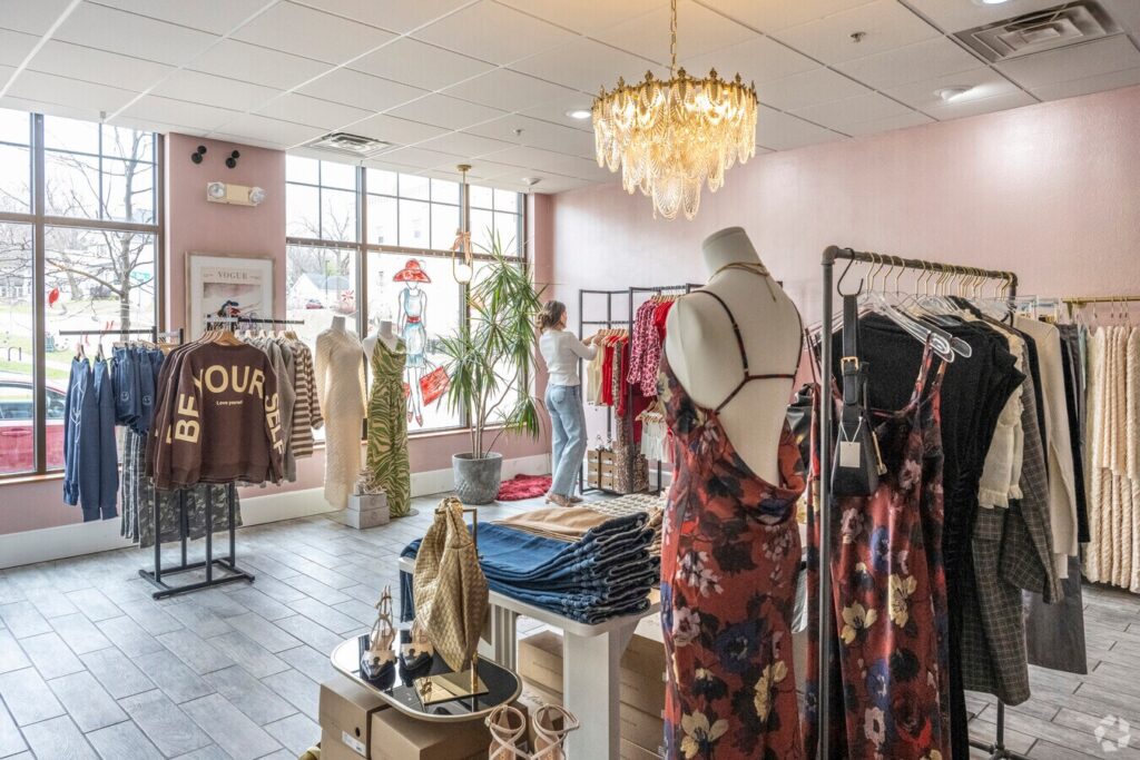 A boutique clothing store with racks of clothes, a mannequin in a floral dress, stacked jeans, and a person arranging items near the window.