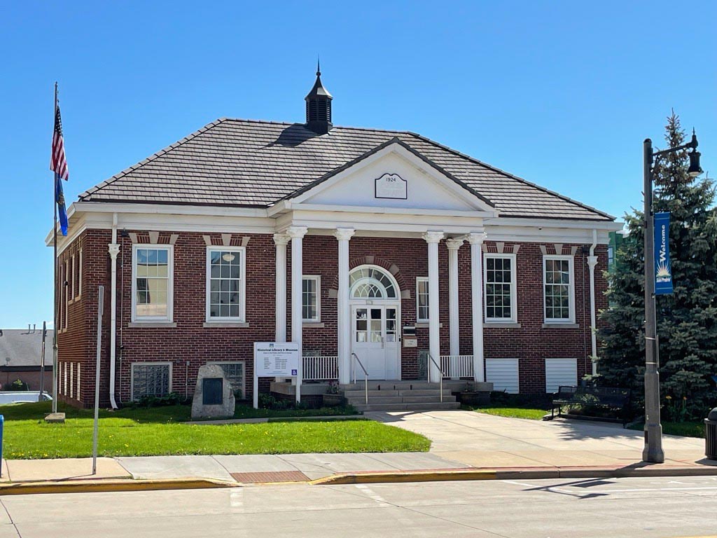 A brick building with white columns and a small cupola, featuring an arched entrance, sits on a sunny street corner with flags and a sign out front.