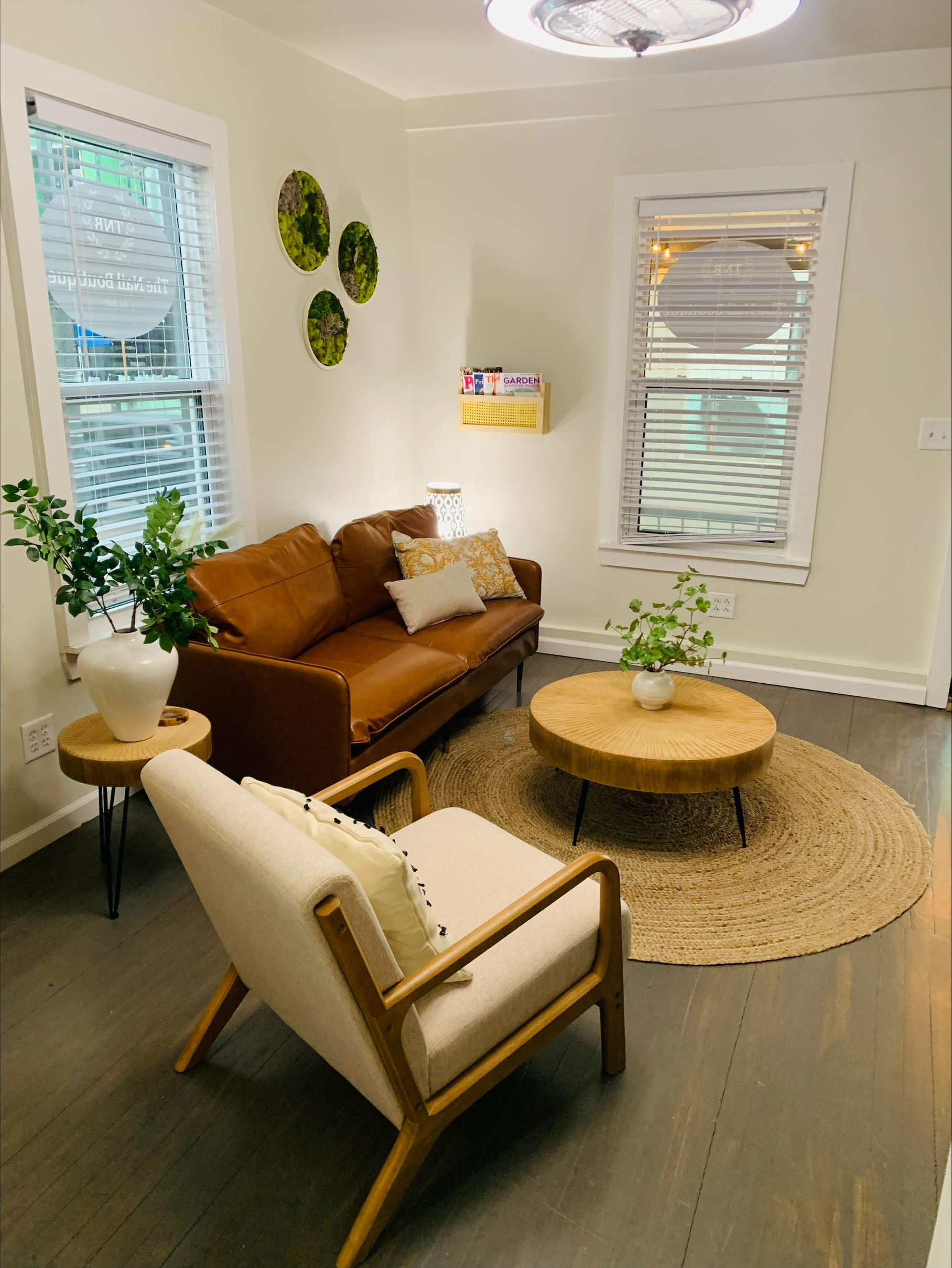 A small living room with a brown leather sofa, a wooden armchair, a round coffee table, potted plants, wall art, and large windows with white blinds.