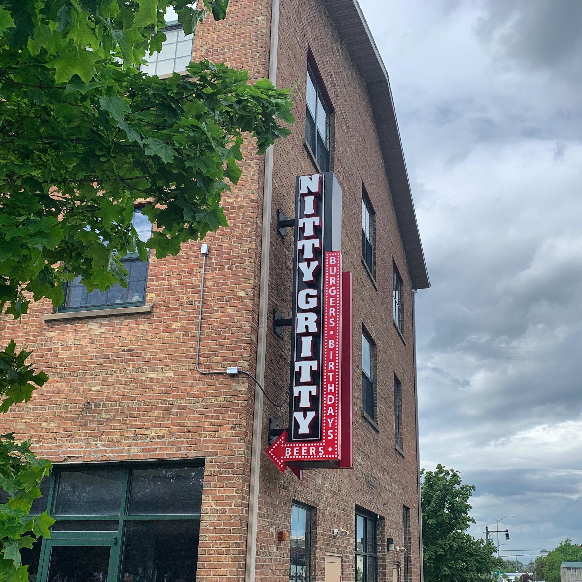 A brick building with a vertical sign reading “Nitty Gritty” and smaller text advertising burgers, birthdays, and beers. The sky is cloudy and a tree is partially visible on the left.