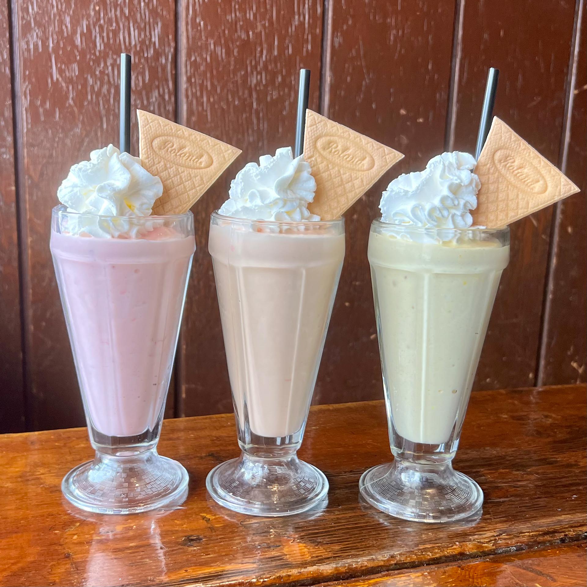 Three milkshakes in tall glasses, each topped with whipped cream, a square wafer biscuit, and a black straw, set on a wooden surface against a wooden wall.