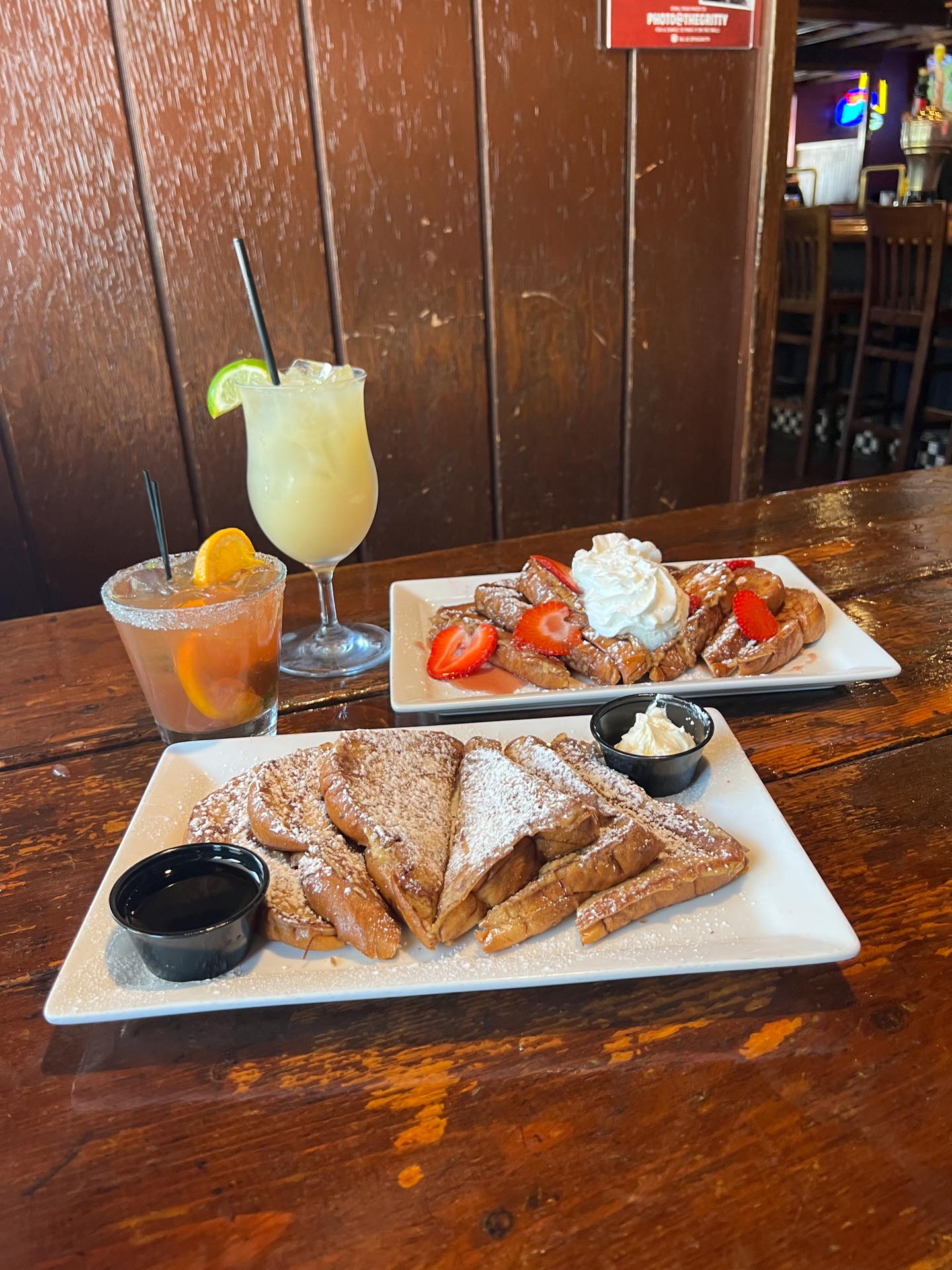 Two plates of French toast with powdered sugar, syrup, and fruit, alongside two cocktails with garnishes, set on a rustic wooden table in a restaurant.